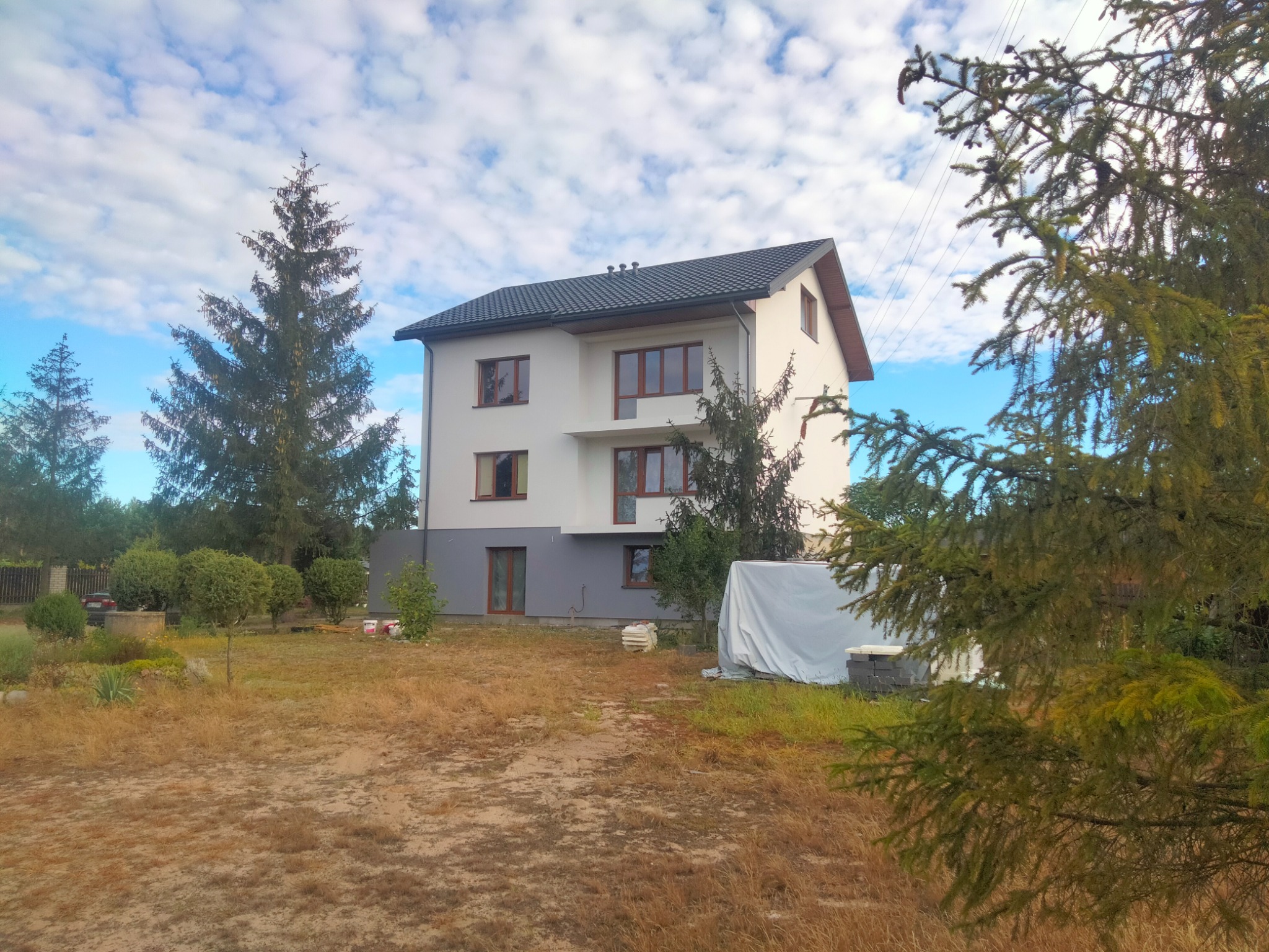 Dwukondygnacyjny dom z białymi ścianami i szarą podstawą, brązowe okna, ciemny dach, otoczony zielenią i uschniętą trawą, z materiałami budowlanymi przykrytymi plandeką z boku.