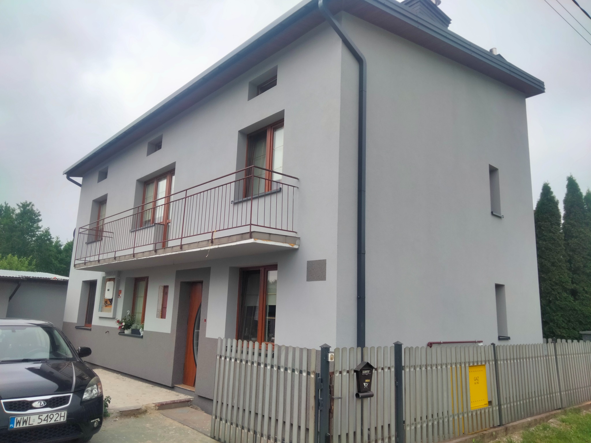 Dwupiętrowy dom z szarą elewacją i drewnianymi oknami, balkon z metalową balustradą, fragment czarnego samochodu na pierwszym planie, widoczna skrzynka na listy przy płocie.