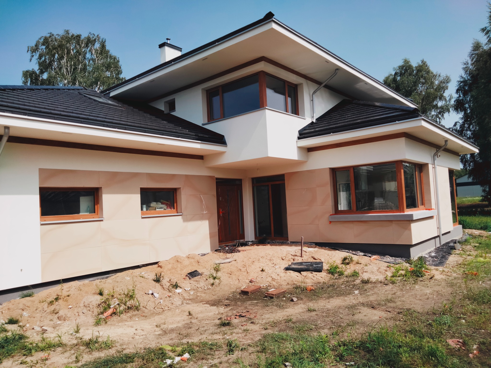 Nowoczesny dom jednorodzinny z jasną elewacją w trakcie prac wykończeniowych, widoczny fragment ogrodu z ziemią i resztkami materiałów budowlanych.