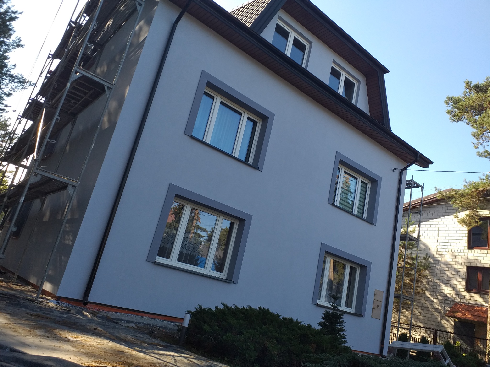 Dwupiętrowy dom z szarą elewacją i ciemnoszarymi obramowaniami okien, częściowo zasłonięty rusztowaniem, z widocznymi refleksami w oknach i krzewami u podstawy.