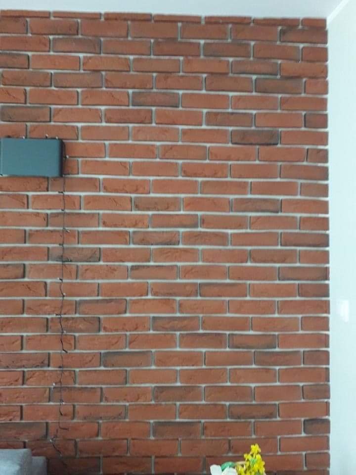 Ściana z cegieł klinkierowych w odcieniach czerwieni i brązu, z widoczną szarą skrzynką elektryczną i delikatnym bukietem kwiatów w dolnym rogu.