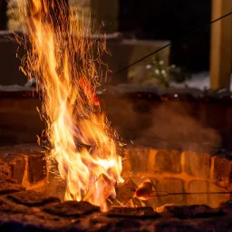 Ognisko w kamiennej obudowie, płomienie i iskry w ciemności, na szpikulcu nad ogniem piecze się kiełbaska.