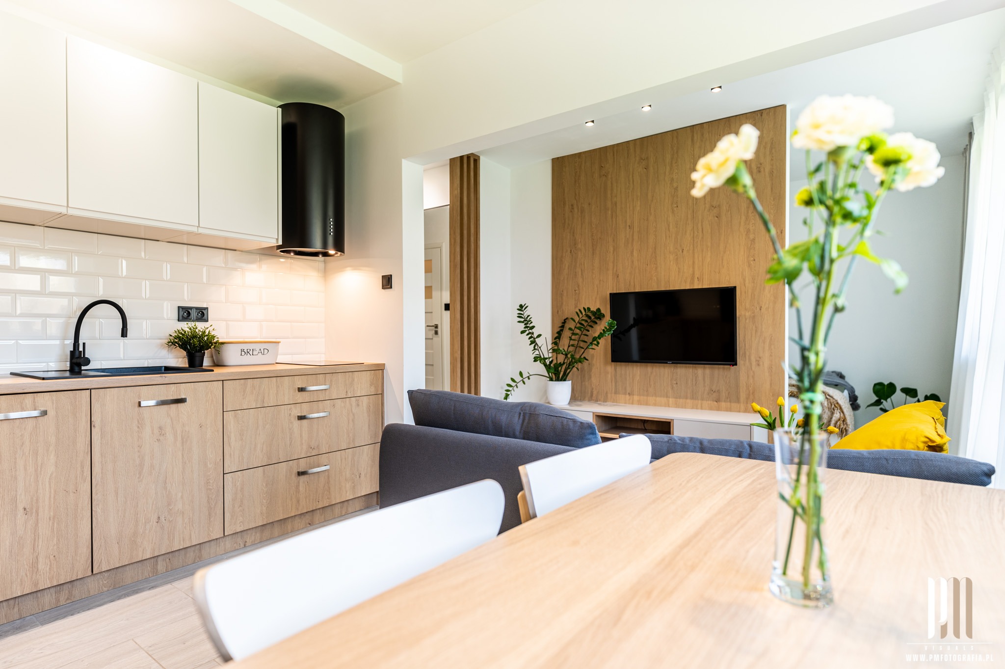 Nowoczesne wnętrze: kuchnia z drewnianymi szafkami i czarną armaturą, połączona z salonem z drewnianą ścianą i telewizorem, kwiaty w wazonie na pierwszym planie.