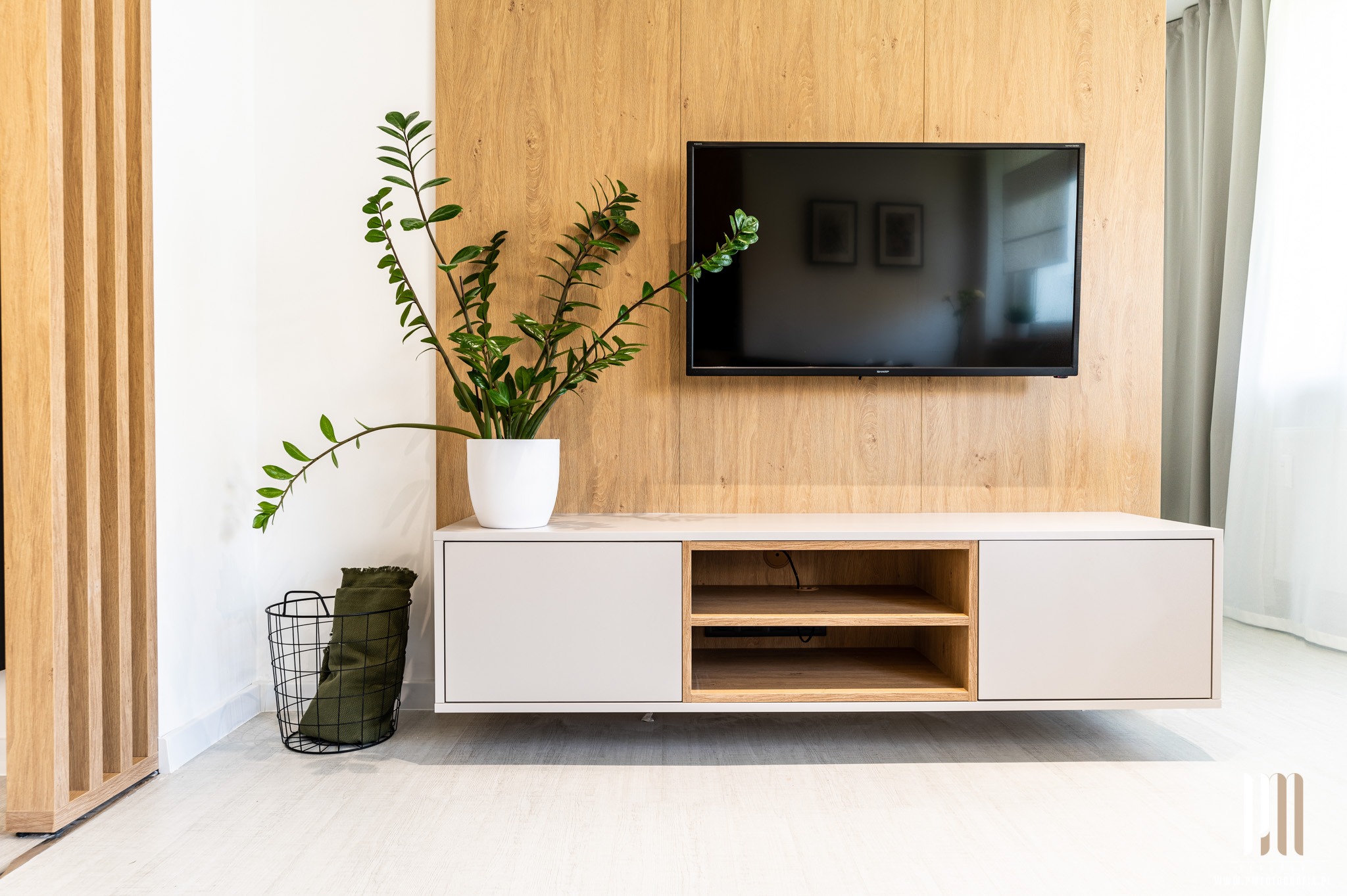 Nowoczesny salon z drewnianą ścianą, wiszącą szafką RTV, telewizorem i zielonym kwiatem w białej doniczce, uzupełniony koszem z pledem.