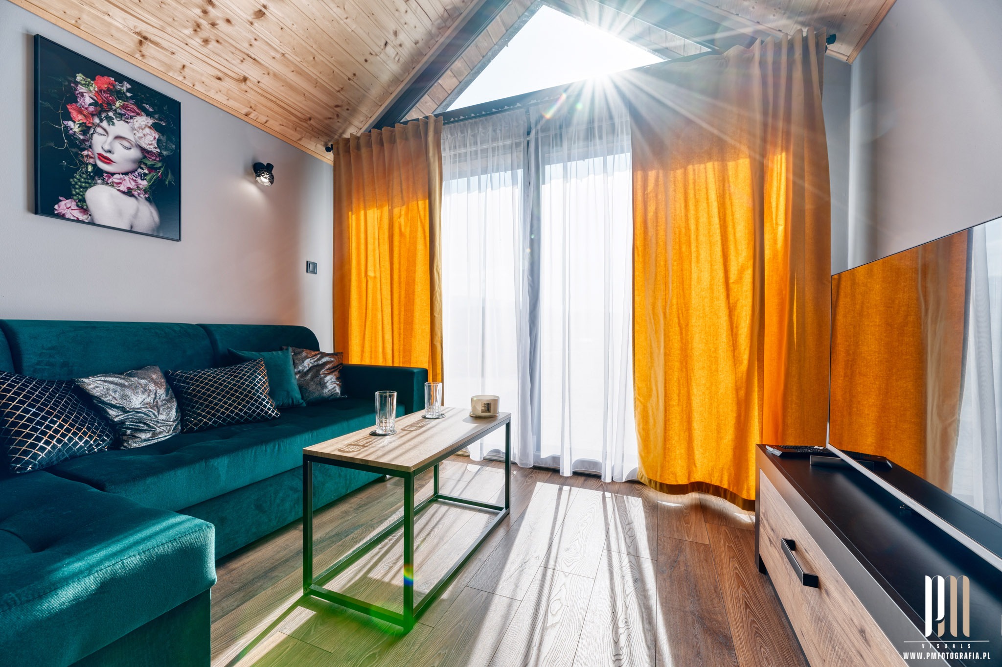 Jasny salon z drewnianym stropem, turkusową sofą, stolikiem kawowym i promieniami słońca wpadającymi przez okno dachowe, z obrazem kobiety z kwiatami na ścianie.