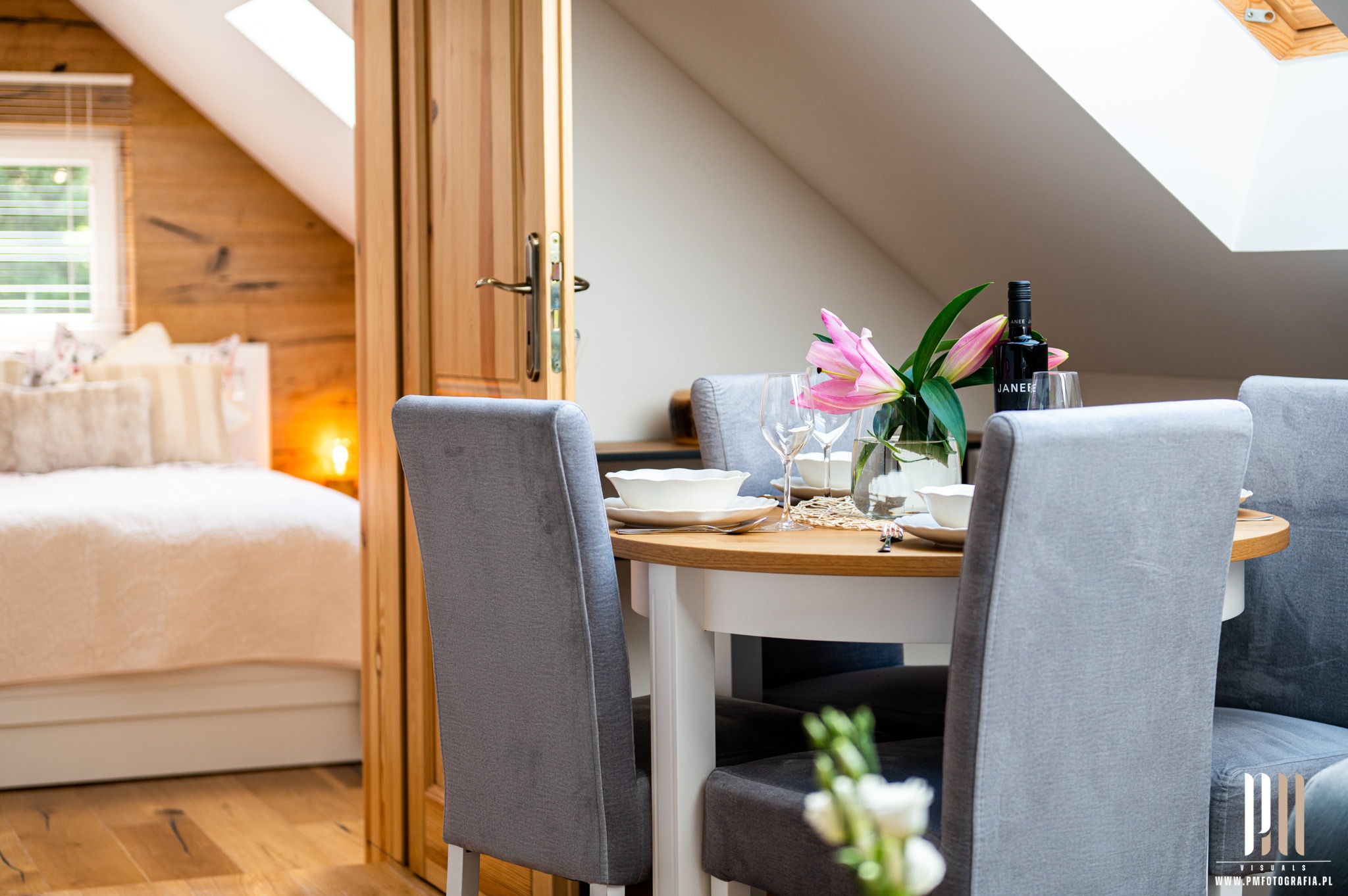 Stół z nakryciem i kwiatami w wazonie, widok na sypialnię z drewnianą boazerią w tle, skos dachu z oknem dachowym