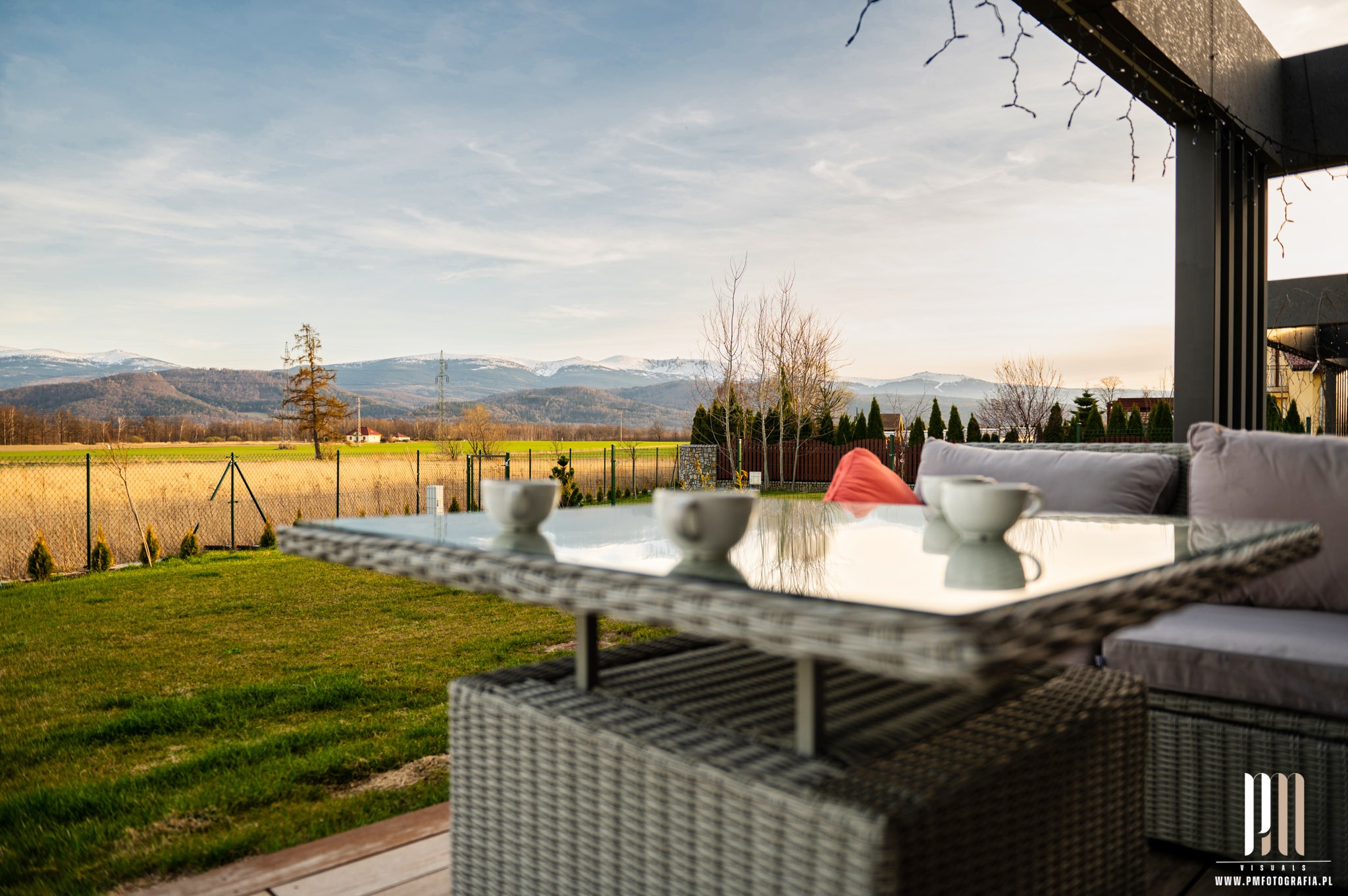 Zewnętrzny salon w Jeleniej Górze z rattanowymi meblami, szklanym stołem, filiżankami i widokiem na góry w tle.