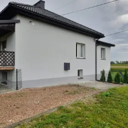 Jasnoszary dom jednorodzinny z ciemną dachówką, widok z boku na elewację, z elementami wykończeniowymi w kolorze szarym, otoczony zielenią i brązowymi kamykami.