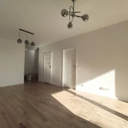 Jasne wnętrze po remoncie: świeżo pomalowane białe ściany, ułożone panele podłogowe, dwie białe drzwi wewnętrzne z przeszkleniami, nowoczesne lampy sufitowe.