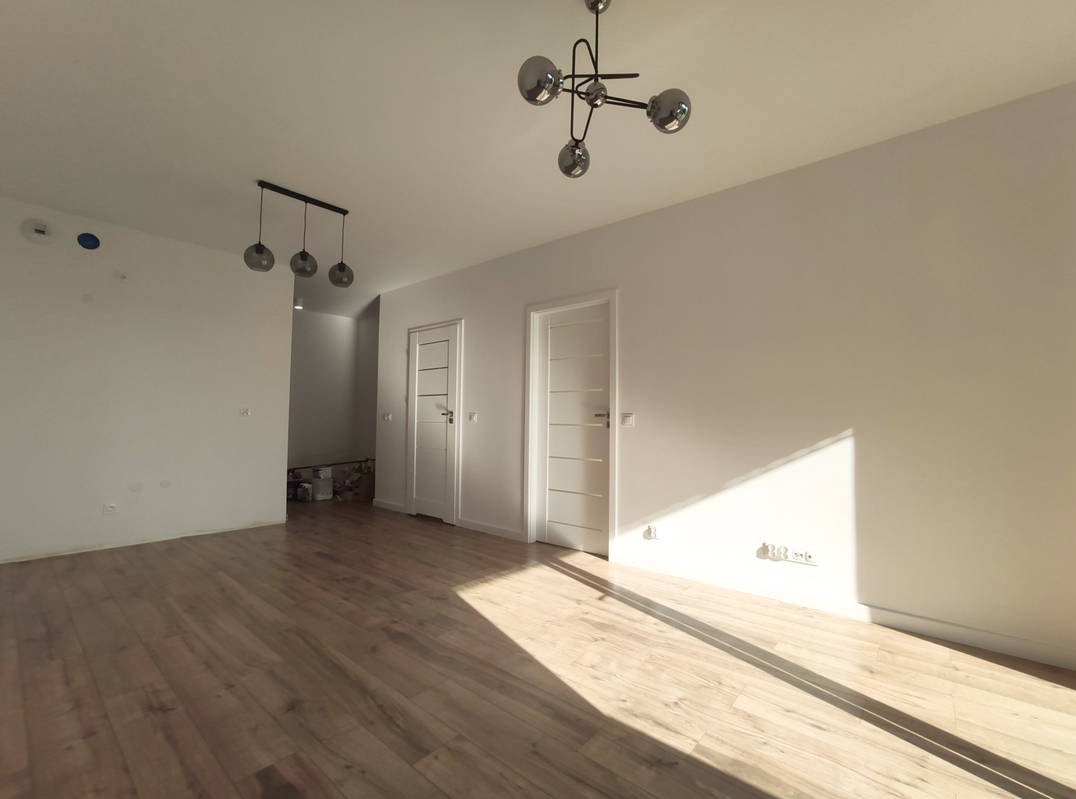 Jasne wnętrze po remoncie: świeżo pomalowane białe ściany, ułożone panele podłogowe, dwie białe drzwi wewnętrzne z przeszkleniami, nowoczesne lampy sufitowe.