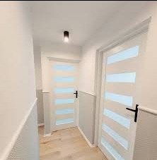 Korytarz po remoncie. Białe ściany z szarym panelem dolnym, jasna drewniana podłoga i dwie białe, nowoczesne drzwi z poziomymi, mlecznymi przeszkleniami oraz czarnymi klamkami.