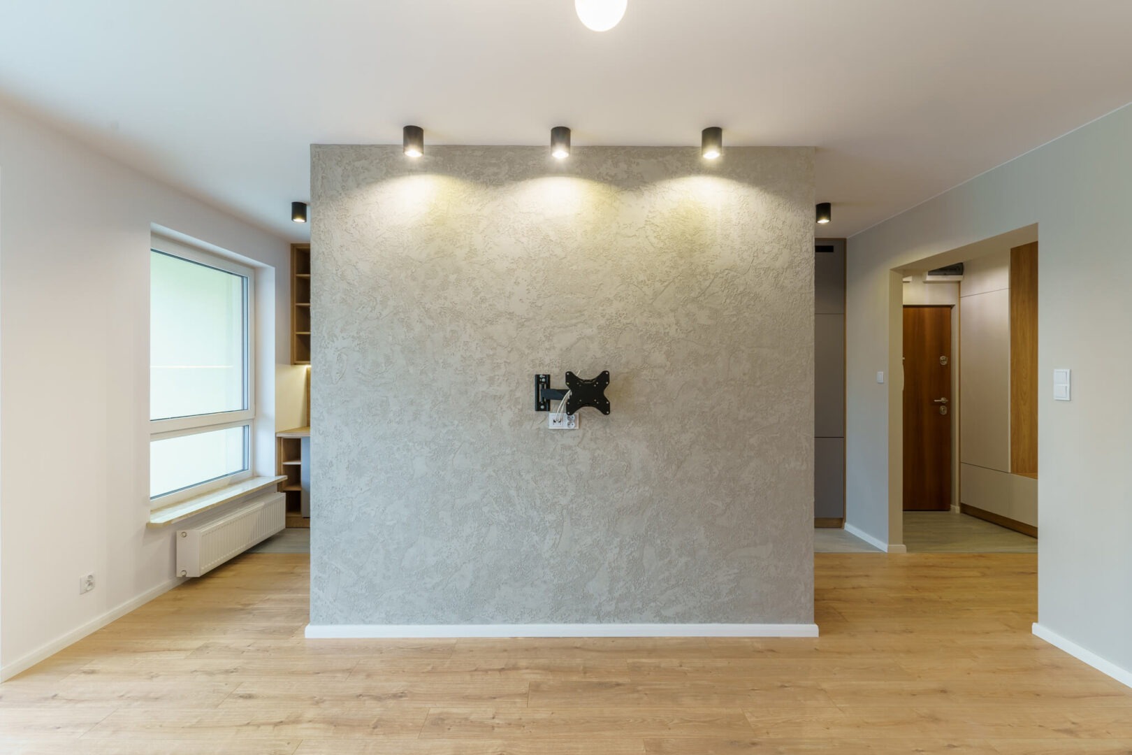 Pusty pokój po remoncie z drewnianą podłogą, ścianą z dekoracyjną fakturą i zamontowanym uchwytem do telewizora.