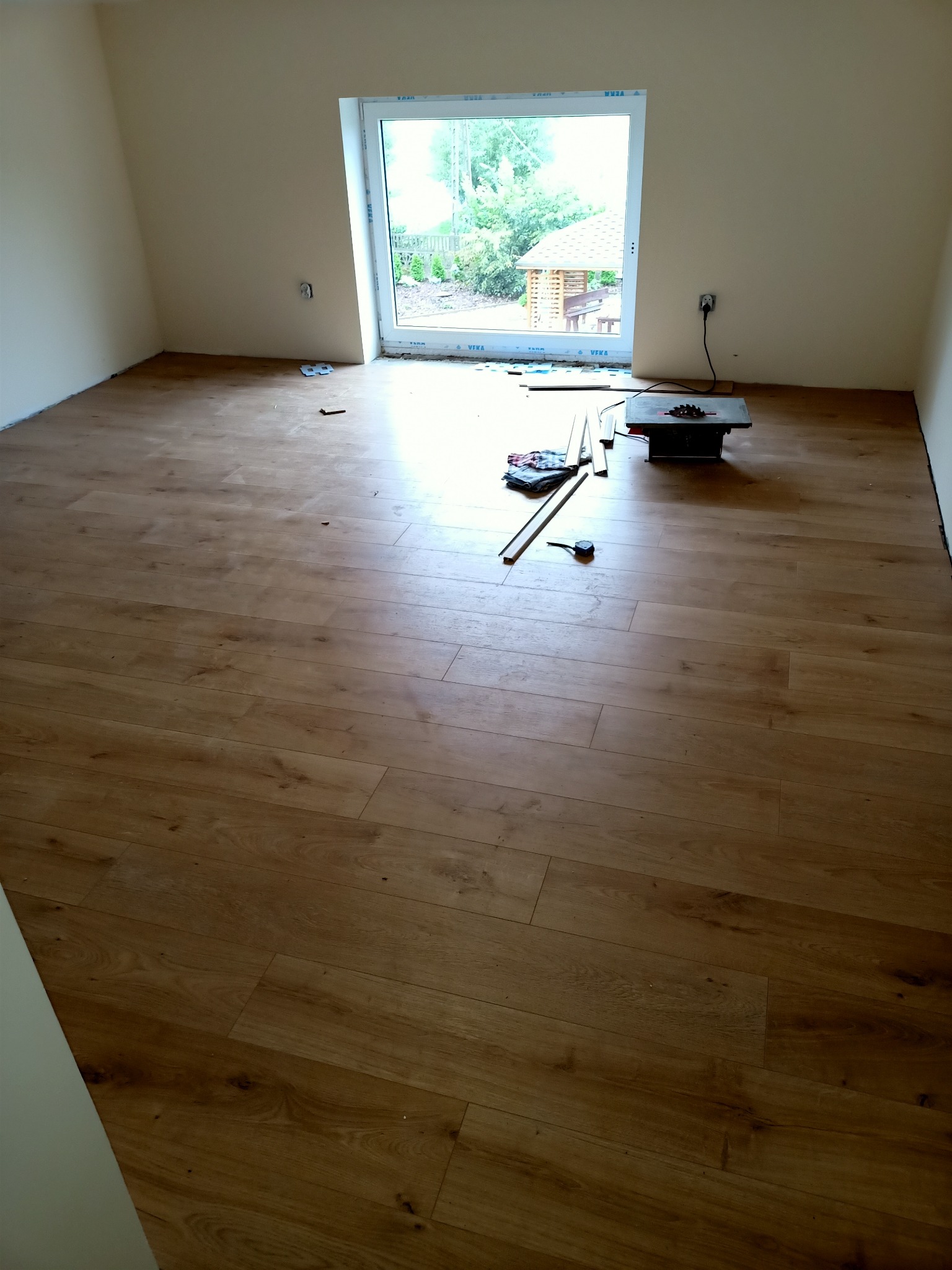 Pokój w trakcie montażu paneli podłogowych z widoczną piłą do cięcia paneli, listwami przypodłogowymi i narzędziami.