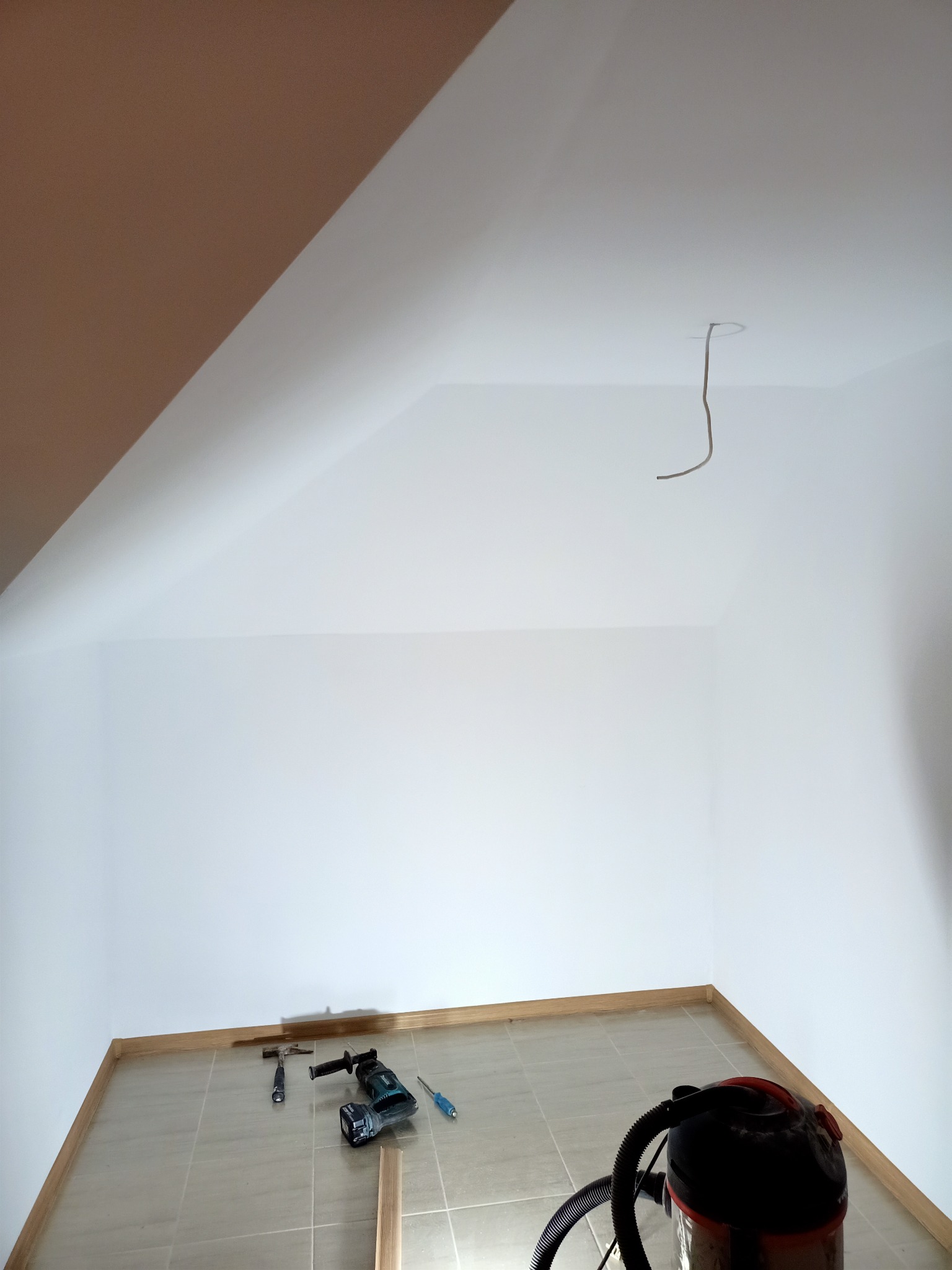 Poddasze w trakcie remontu: świeżo pomalowane białe ściany, listwy przypodłogowe, narzędzia (wiertarka, młotek, śrubokręt) i odkurzacz przemysłowy na podłodze z płytek.