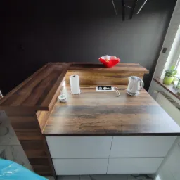 Blat kuchenny z drewna o ciemnym odcieniu, z widocznymi białymi szafkami poniżej, wyposażony w gniazda elektryczne, czajnik, pojemnik na papierowe ręczniki i czerwoną misę na owoce na tle czarnej...