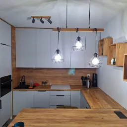 Nowoczesna kuchnia z szarymi frontami szafek, drewnianym blatem i dekoracyjną ścianą nad blatem, oświetlona trzema wiszącymi lampami w stylu industrialnym oraz dodatkowymi reflektorami.