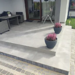 Wykończony taras z szarych płytek ceramicznych, dwustopniowe schody, meble ogrodowe z rattanu i donice z wrzosem, widok na kostkę brukową przed domem.