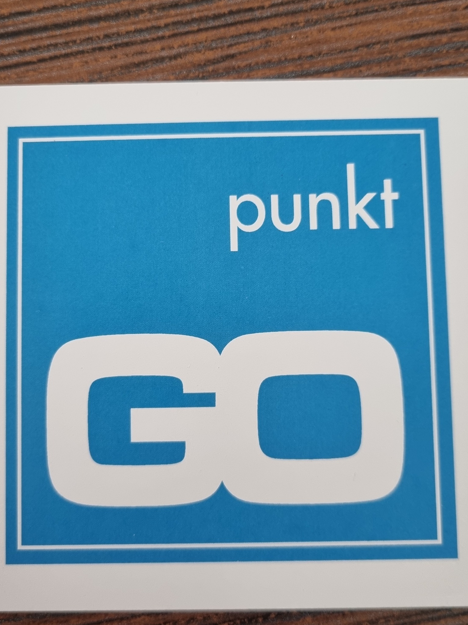 Białe logo 'punkt GO' na błękitnym tle w białej ramce, leżące na drewnianym blacie.