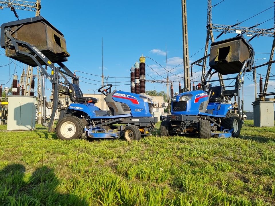 Dwa niebieskie traktorki ISEKI z podnośnikami do śniegu na tle stacji transformatorowej, trawa i błękitne niebo.