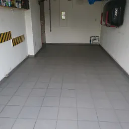 Wygląd świeżo wykafelkowanej podłogi w garażu, z szarymi, kwadratowymi płytkami ułożonymi r&oacute;wno, widoczne rury instalacji wodnej przy ścianie, p&oacute;łki z farbami i akcesoriami na ścianie po lewej,...