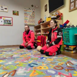Dwoje ratowników medycznych w strojach ochronnych klęczy na kolorowym dywanie z literami w sali zabaw, prawdopodobnie podczas szkolenia z pierwszej pomocy dla dzieci, widoczna apteczka i zabawki.