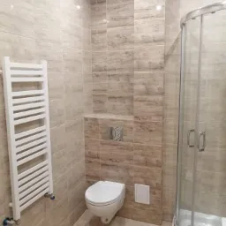 Wykończona łazienka z beżowymi płytkami imitującymi drewno, wisząca toaleta, grzejnik drabinkowy i kabina prysznicowa z chromowanymi detalami.