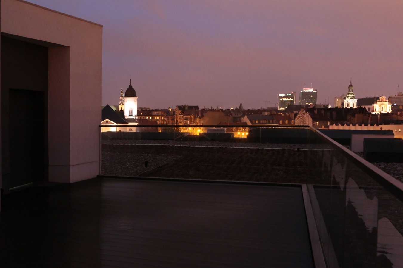 Nocny widok z balkonu apartamentu na panoramę miasta z oświetlonymi budynkami, widoczna szklana balustrada i fragment elewacji budynku.