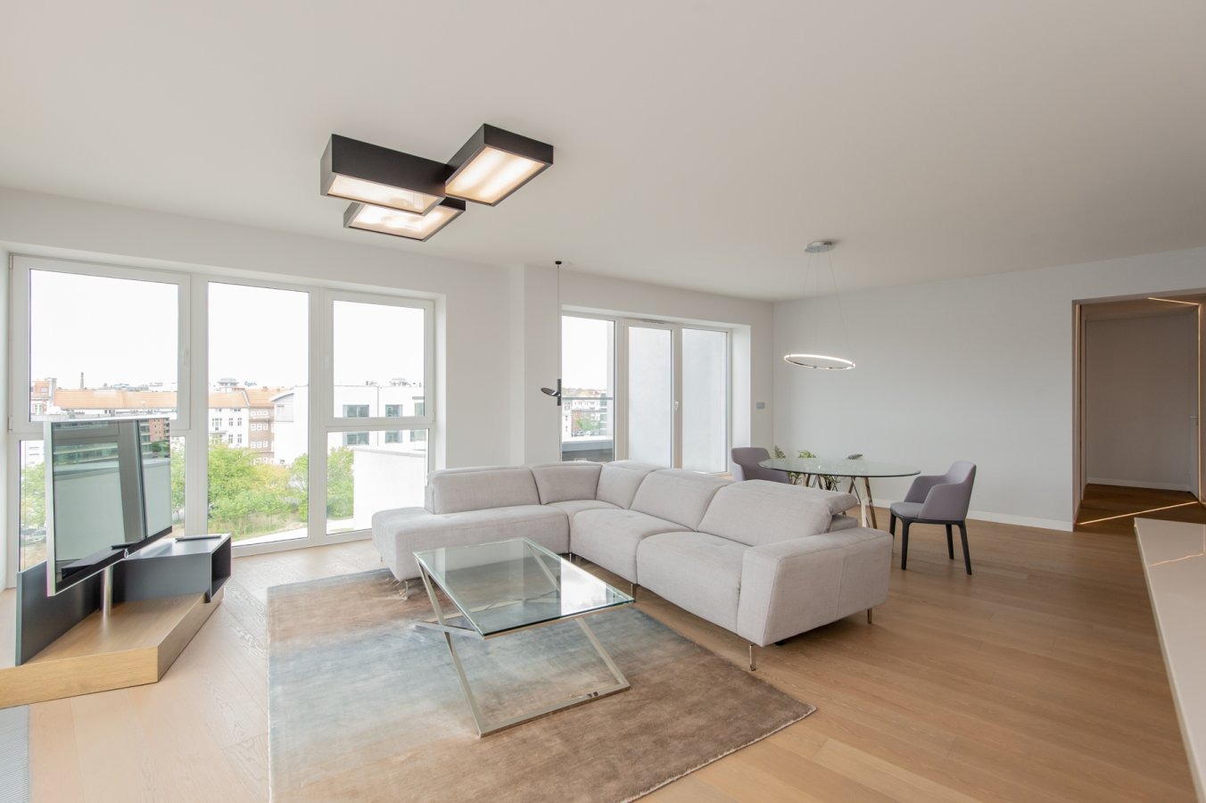 Jasne, przestronne wnętrze salonu z narożną sofą, szklanym stolikiem kawowym na wzorzystym dywanie, telewizorem i widokiem na miasto przez duże okna, oświetlone nowoczesnymi lampami sufitowymi.