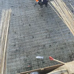 Pracownik w pomarańczowej kurtce układa zbrojenie na przygotowanym fundamencie pod budowę domu, widoczne stalowe pręty i siatka zbrojeniowa.