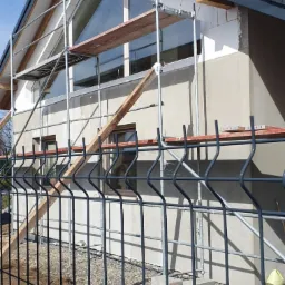 Dom w trakcie prac elewacyjnych, widoczne rusztowania, fragment dachu z ciemną dach&oacute;wką, ściany częściowo ocieplone, taczka i narzędzia na pierwszym planie za ogrodzeniem.