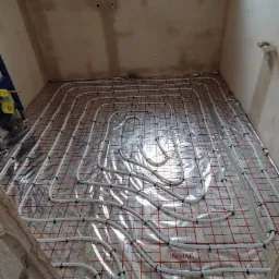 Instalacja ogrzewania podłogowego z rur na folii z siatką w pomieszczeniu w trakcie remontu, widoczne okno dachowe i surowe ściany.