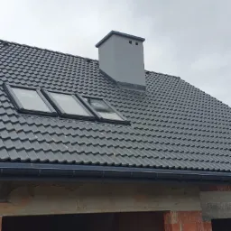 Nowy dach z szarej dachówki z trzema oknami dachowymi i szarym kominem na tle pochmurnego nieba, widoczny fragment muru z czerwonej cegły w stanie surowym.