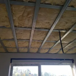 Poddasze w trakcie ocieplania wełną mineralną, widoczne metalowe profile konstrukcyjne sufitu podwieszanego i fragment okna z widokiem na drzewa.