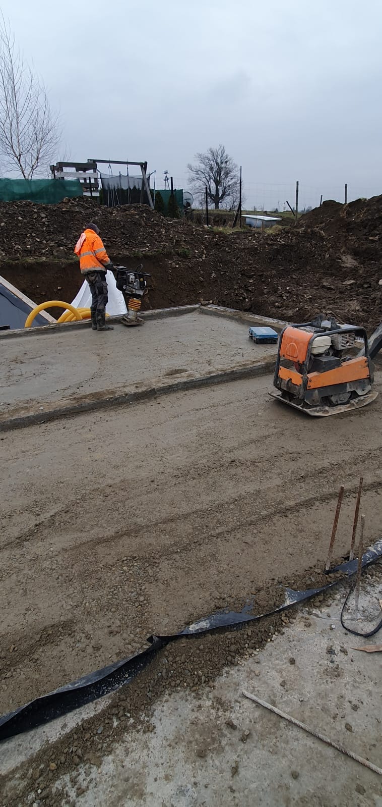 Pracownik w pomarańczowej kurtce zagęszcza podłoże pod fundamenty domu za pomocą zagęszczarki w pochmurny dzień. Na drugim planie widoczna wykopana dziura w ziemi.