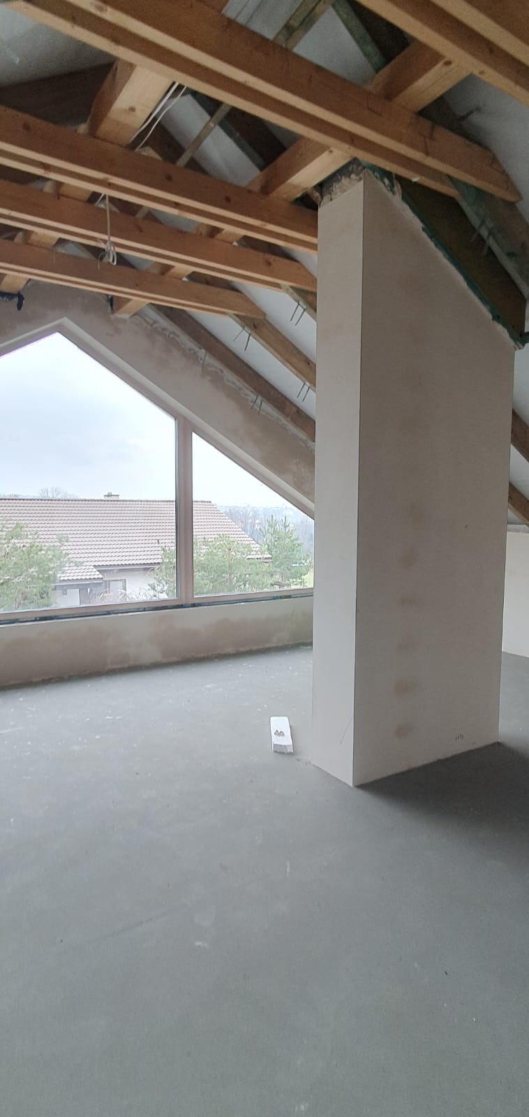 Poddasze w trakcie adaptacji: widoczne drewniane krokwie, okno dachowe z widokiem na dom i drzewa, oraz fragment ściany z pionowym słupem obłożonym płytami gipsowo-kartonowymi.