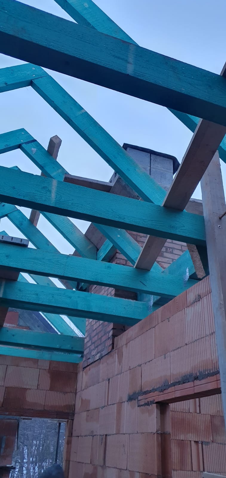 Niebieskie krokwie dachowe w trakcie montażu na murowanym domu z widocznym kominem, surowy stan.