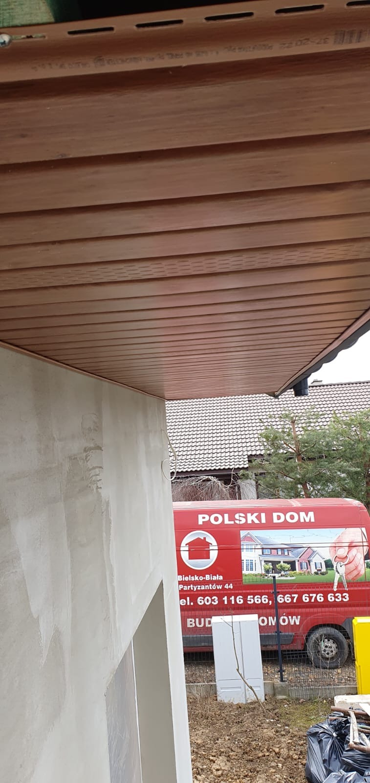 Wykończone drewnopodobne podbitki dachowe z widoczną nowo otynkowaną ścianą budynku i samochodem firmowym w tle.