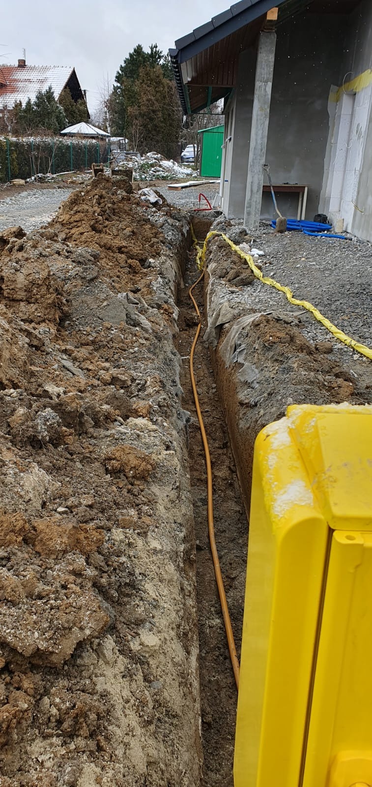 Wykop w ziemi z żółtą rurą i taśmą ostrzegawczą, widoczna skrzynka przyłączeniowa, w tle budynek w trakcie budowy.