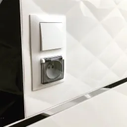 Biały włącznik światła i gniazdko elektryczne z przezroczystą klapką ochronną IP44, zamontowane na białej, wzorzystej ścianie z czarnymi akcentami.