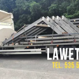 Biała laweta z napisem 'Pomoc Drogowa' przewozi metalowe konstrukcje w kształcie trójkątów, zabezpieczone łańcuchami.