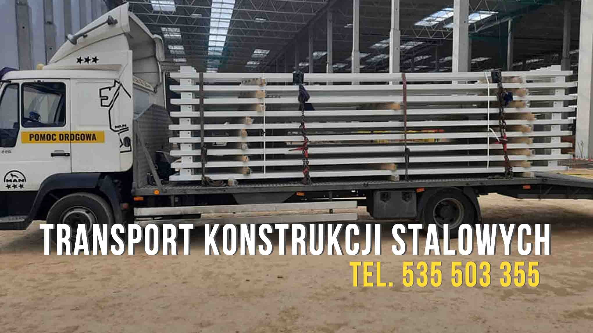 Ciężarówka z platformą załadowana białymi elementami konstrukcji stalowych, zabezpieczonymi łańcuchami, na tle hali przemysłowej.