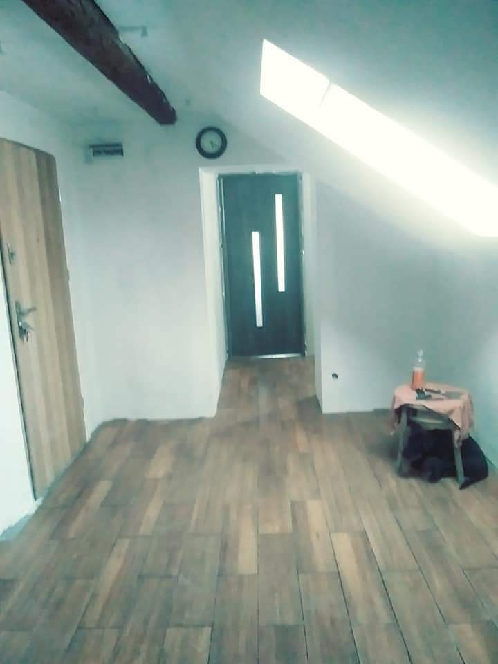 Wykończone poddasze z drewnopodobną podłogą, białe ściany, okno dachowe i widoczne elementy konstrukcyjne, takie jak belka stropowa.