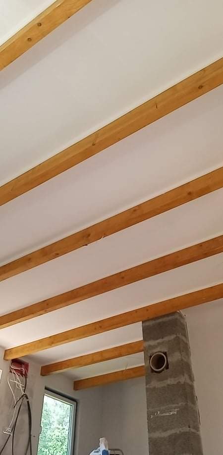 Biały sufit z widocznymi drewnianymi belkami, surowy komin z otworem na rurę, okno i instalacja elektryczna w trakcie montażu.