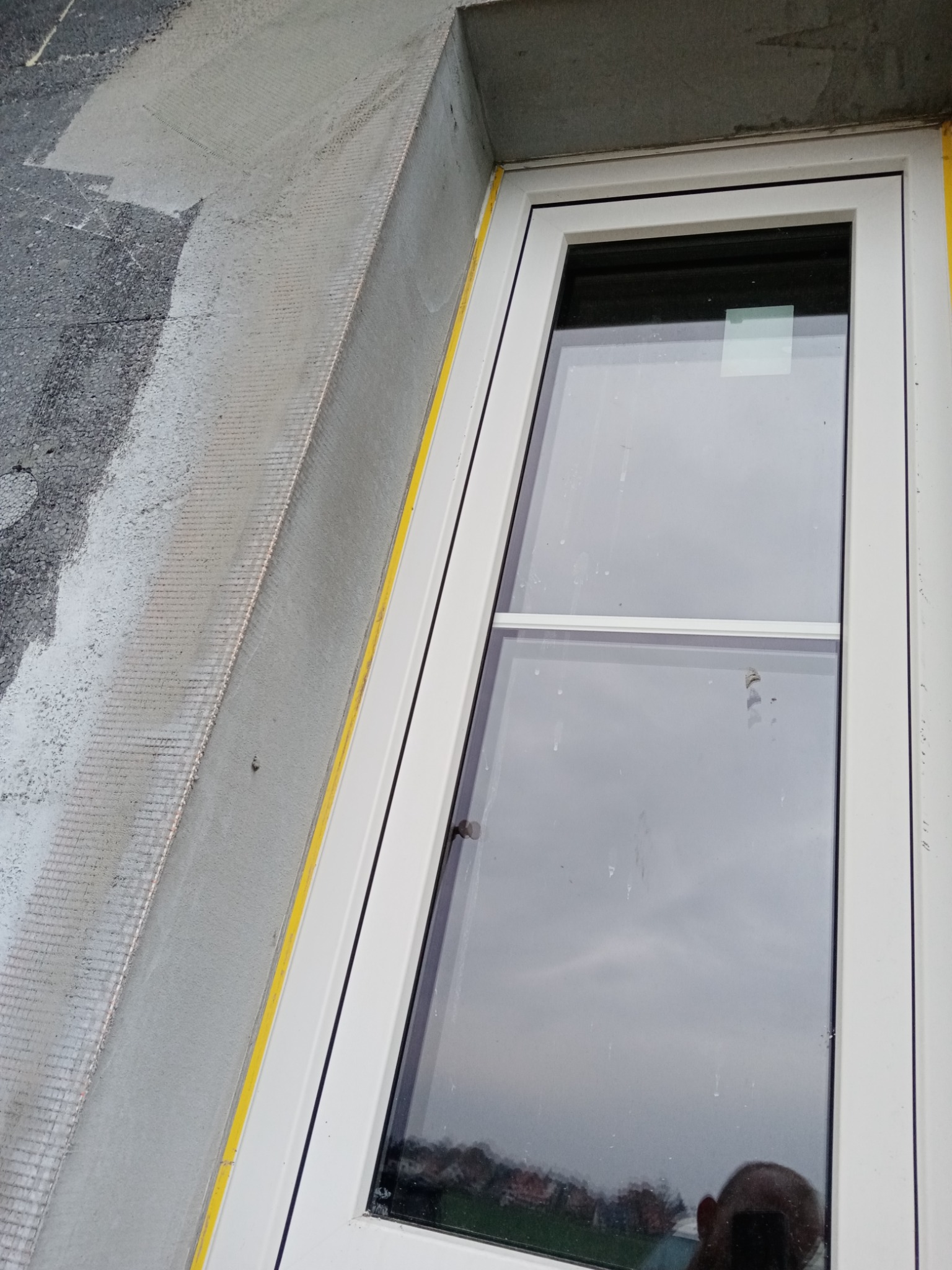 Nowe białe okno PCV z żółtą taśmą malarską na styku z surową elewacją z siatką i częściowo nałożonym tynkiem.