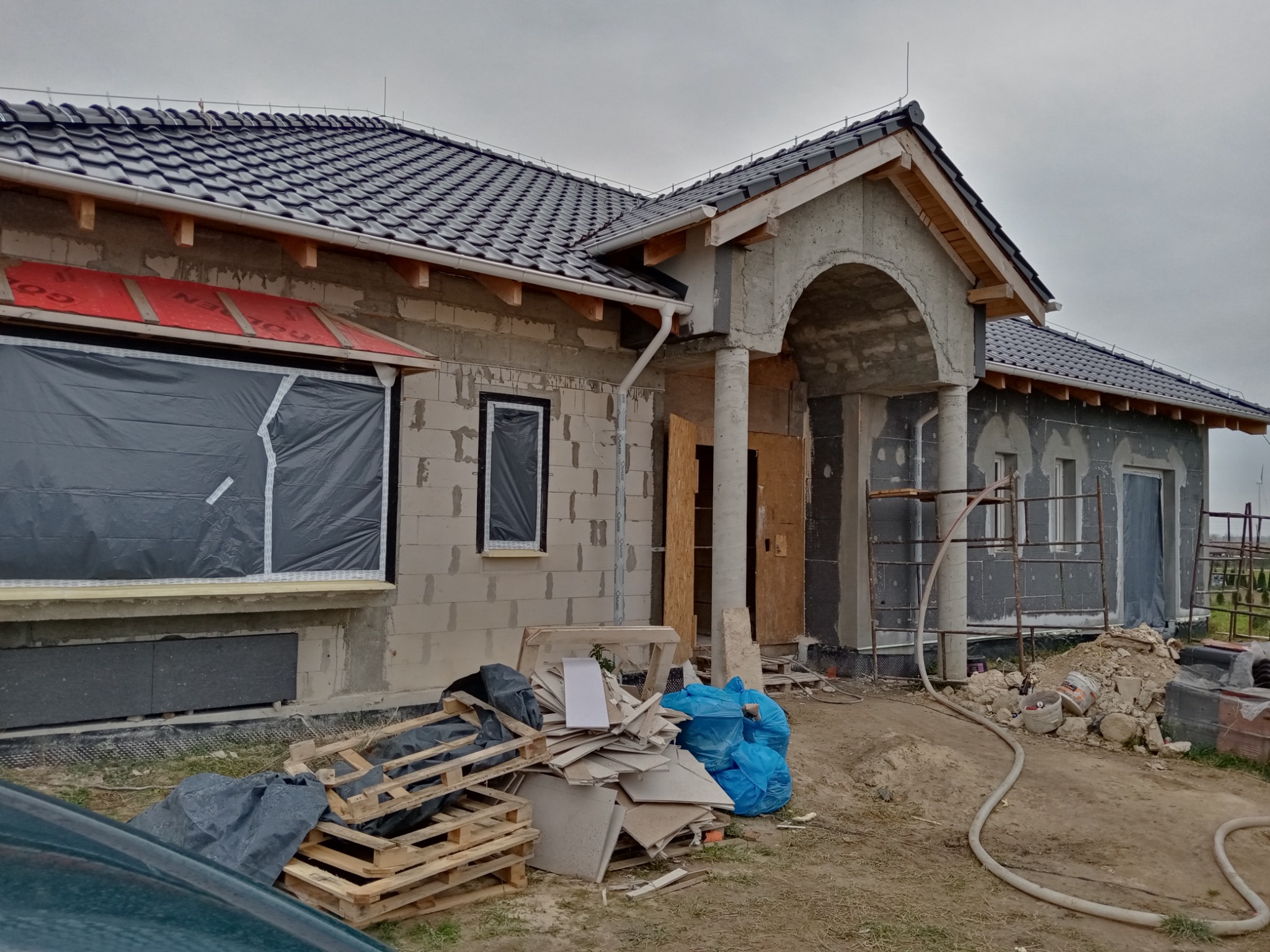 Dom w trakcie budowy z czarną dachówką, widoczne okna zabezpieczone folią, elewacja w stanie surowym, materiały budowlane na paletach i workach