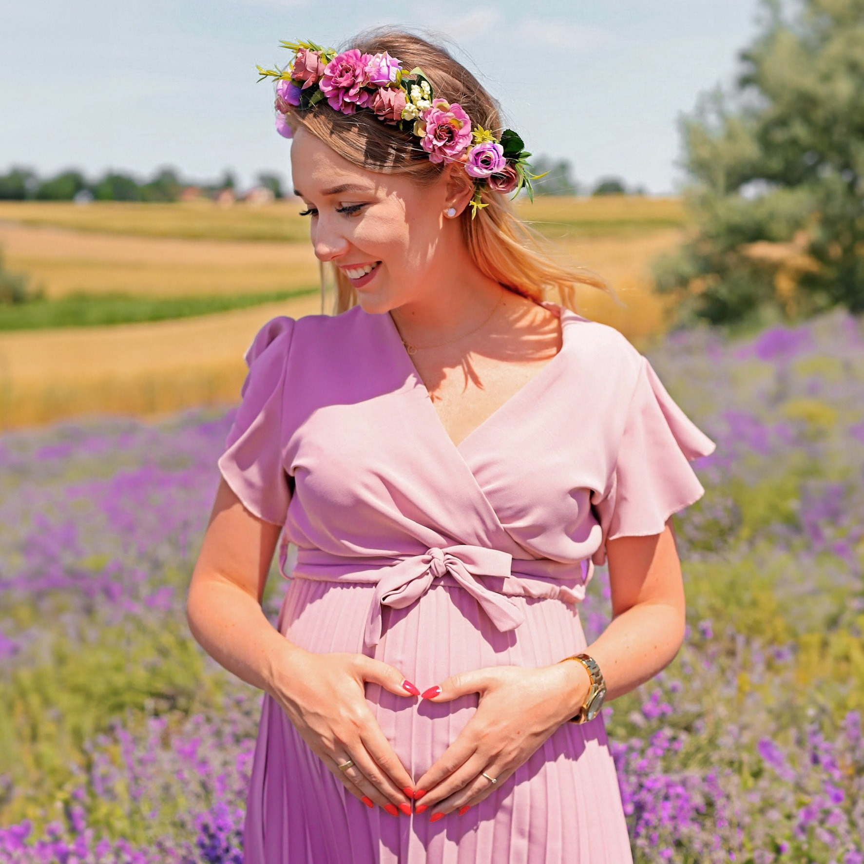 Uśmiechnięta, ciężarna blondynka w różowej sukience i kwiatowym wianku, dłońmi formuje serce na brzuchu, lawendowe pole w tle.