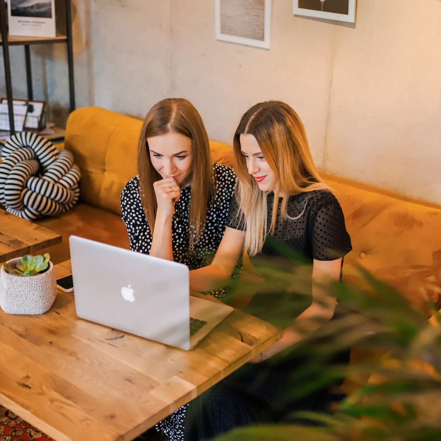 Dwie kobiety w kawiarni, pochylone nad laptopem na drewnianym stole, analizują projekt strony internetowej, widoczny sukulent w doniczce i fragment wzorzystego dywanu.