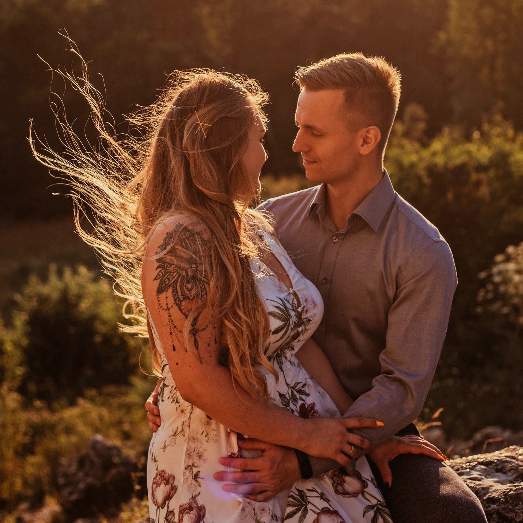 Para zakochanych, kobieta w kwiecistej sukience z tatuażem na ramieniu, mężczyzna w szarej koszuli, obejmują się na tle rozświetlonej słońcem roślinności, włosy kobiety rozwiane przez wiatr.