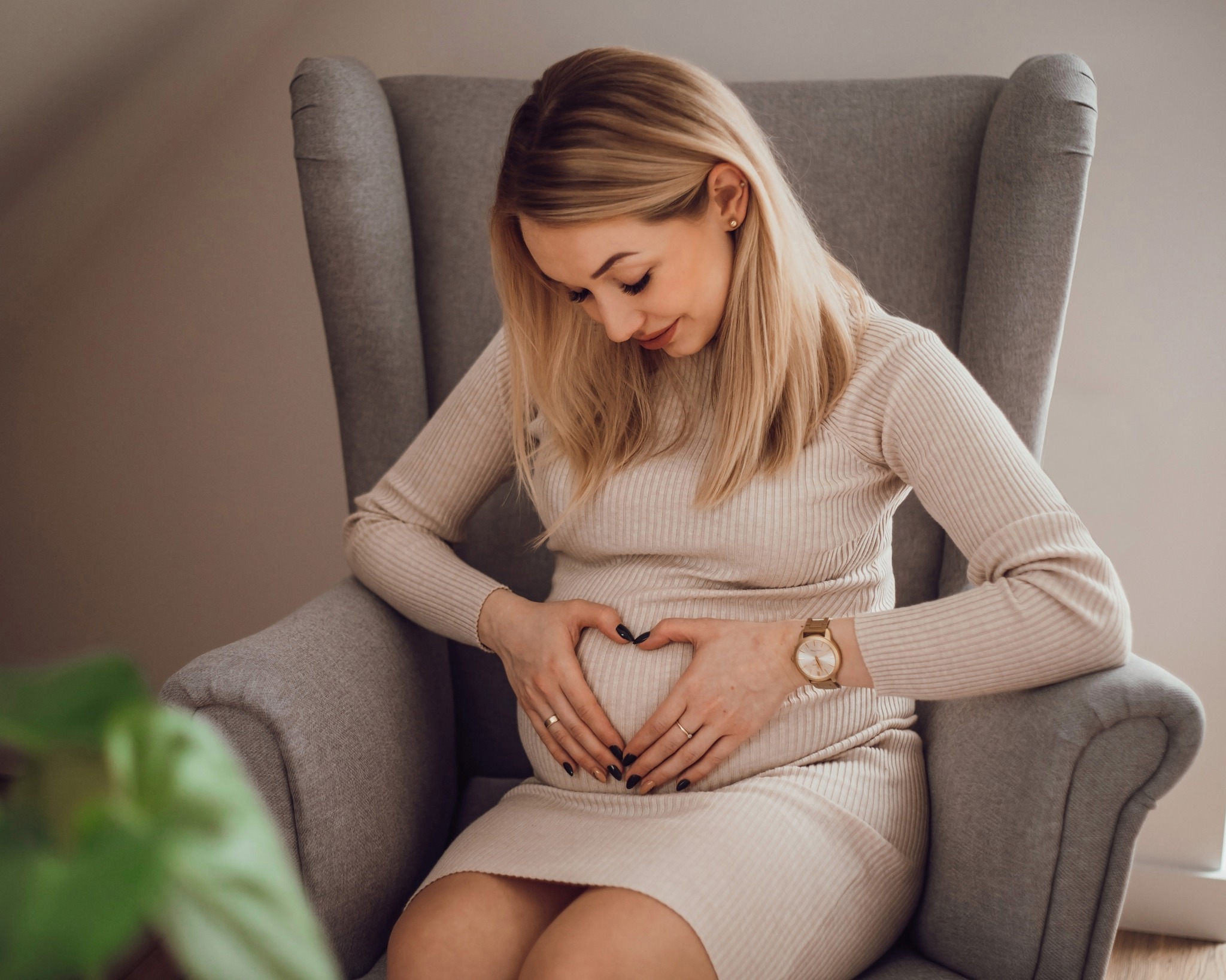 Kobieta w beżowej sukience siedzi w szarym fotelu, dłońmi formuje serce na brzuchu, na nadgarstku złoty zegarek.