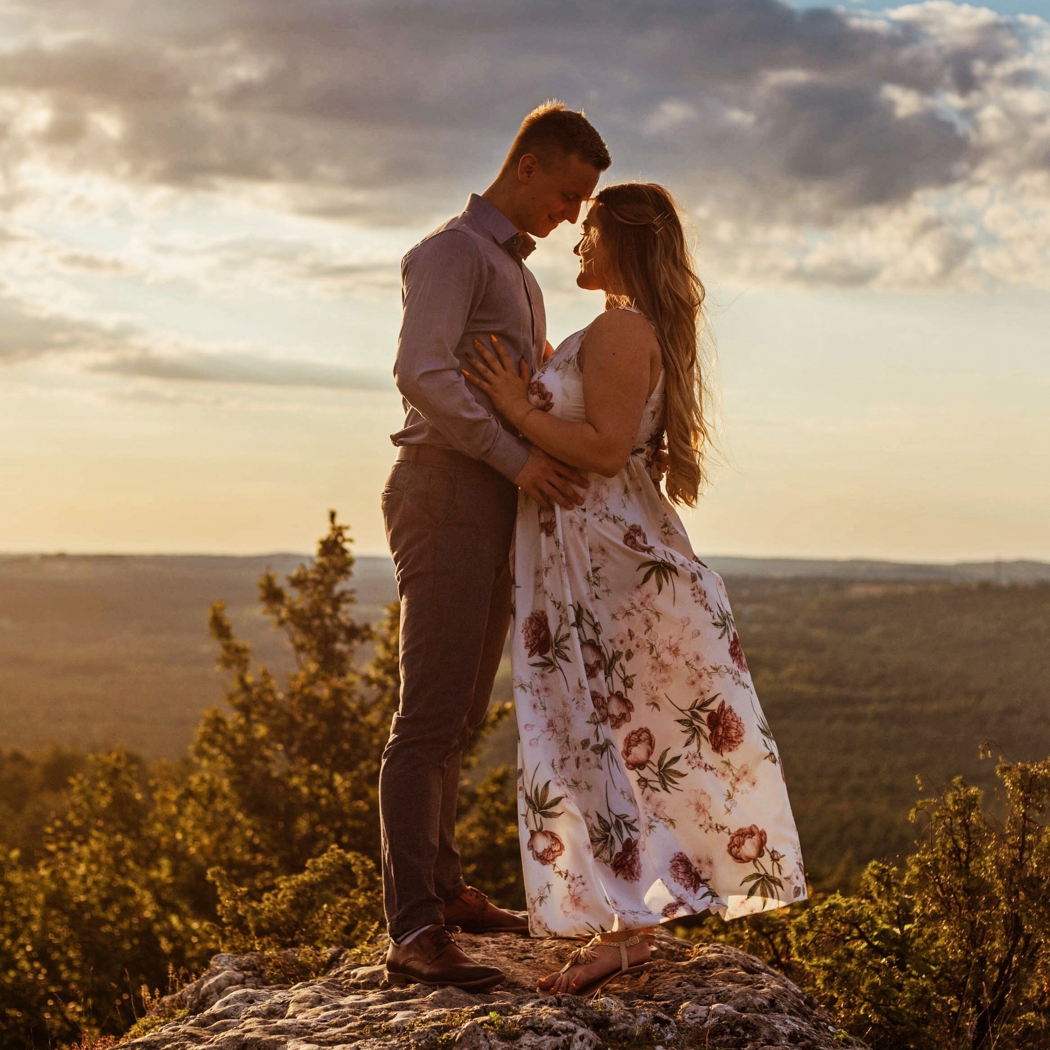 Para zakochanych stoi na szczycie skały, tuląc się do siebie na tle zachodzącego słońca i malowniczej panoramy.